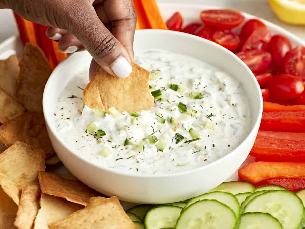 Side view of tzatziki sauce in a white serving dish with a pita chip being dipped in the sauce.