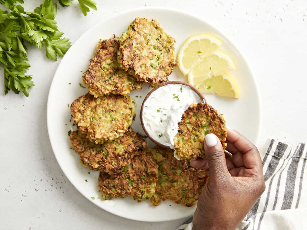 Overhead view of zucchini fritters on a platter served with a hand dipping one fritter in tzatziki sauce.