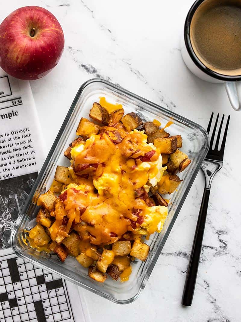 One Country Breakfast Bowl meal prep container on a table with a morning paper, mug of coffee, and an apple.