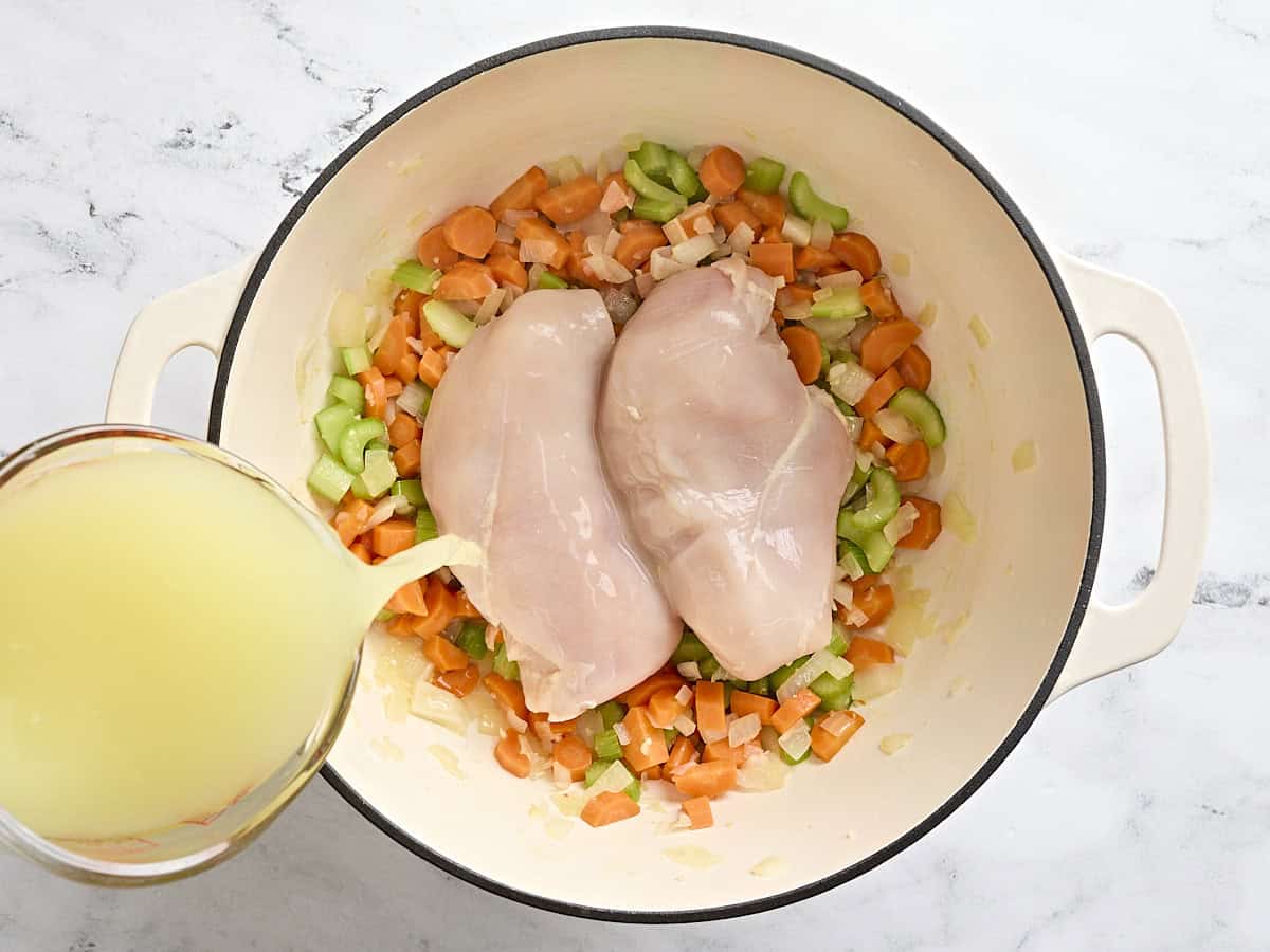 Raw chicken added to sauteed mirepoix in a sauce pot, with chicken broth being poured on top.