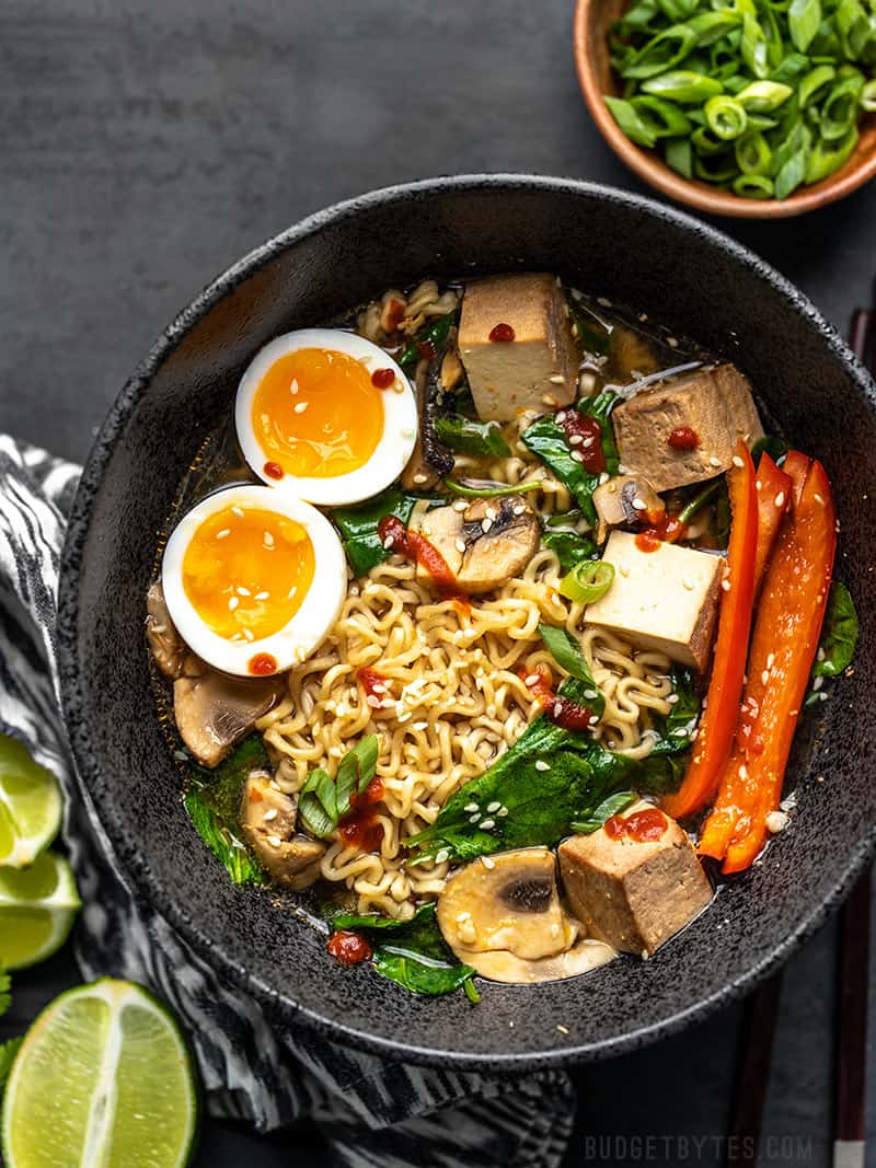 A big bowl of ramen with a soft boiled egg, tofu cubes, spinach, bell pepper, and sliced green onion on the side.
