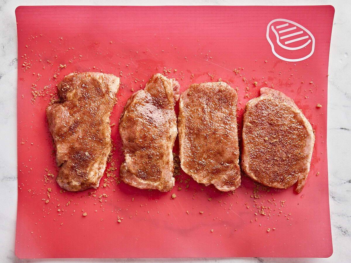 Four pork chops, covered in seasonings on a meat preparation mat.