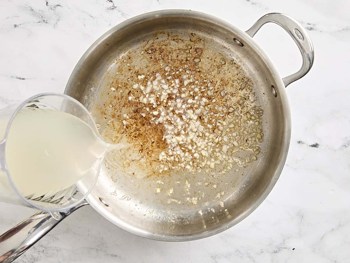 Reserved pasta water being poured into a skillet with minced garlic.