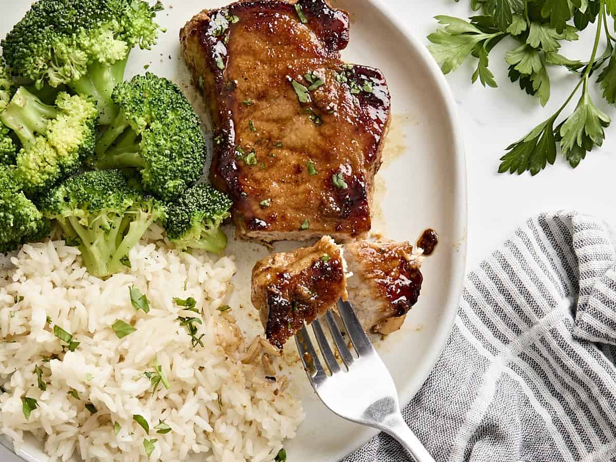 A fork taking a slice of glazed pork chops from a plate with broccoli and rice.