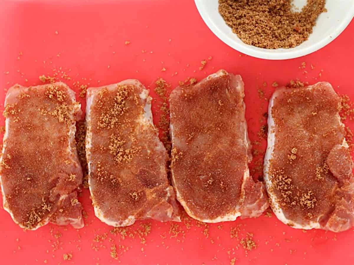 Raw pork chops coated in a spice rub on a red cutting board.