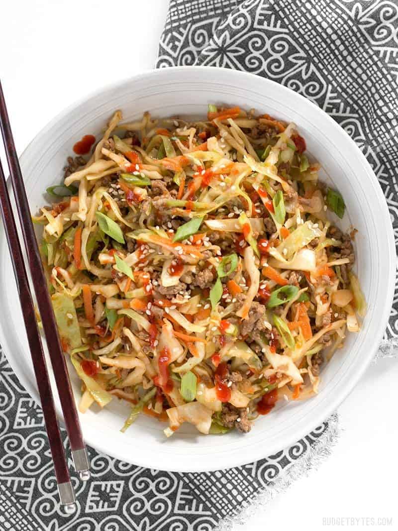 A bowl of Beef and Cabbage Stir Fry topped with sriracha, chopsticks on the side