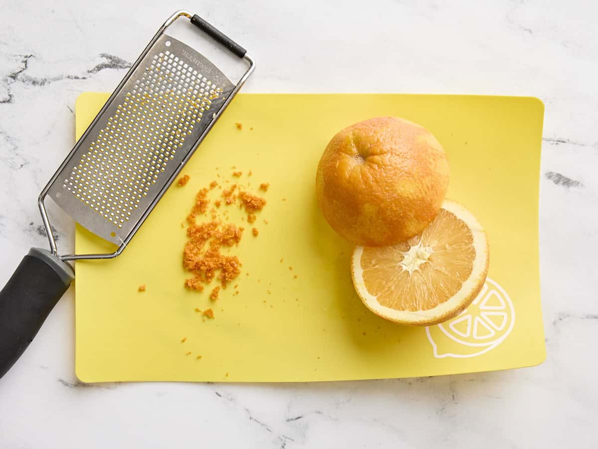 Two orange halves on a cutting board with orange zest and a zester.