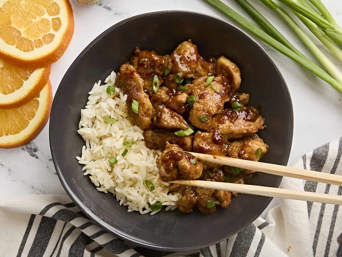 Overhead view of orange chicken in a bowl with rice, with chopsticks taking a piece of chicken.