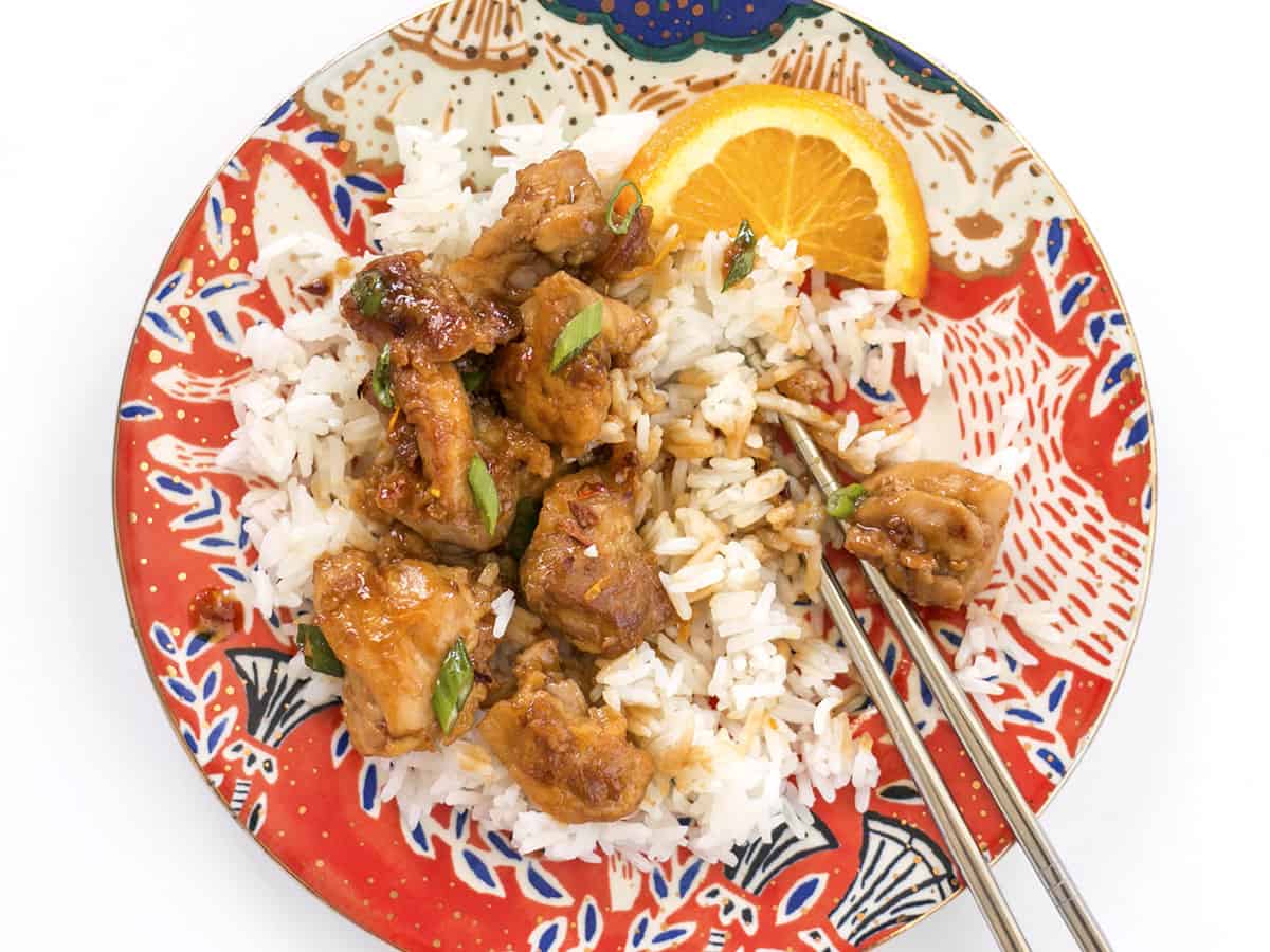 A plate of rice and orange chicken in the process of being eaten with silver chopsticks.