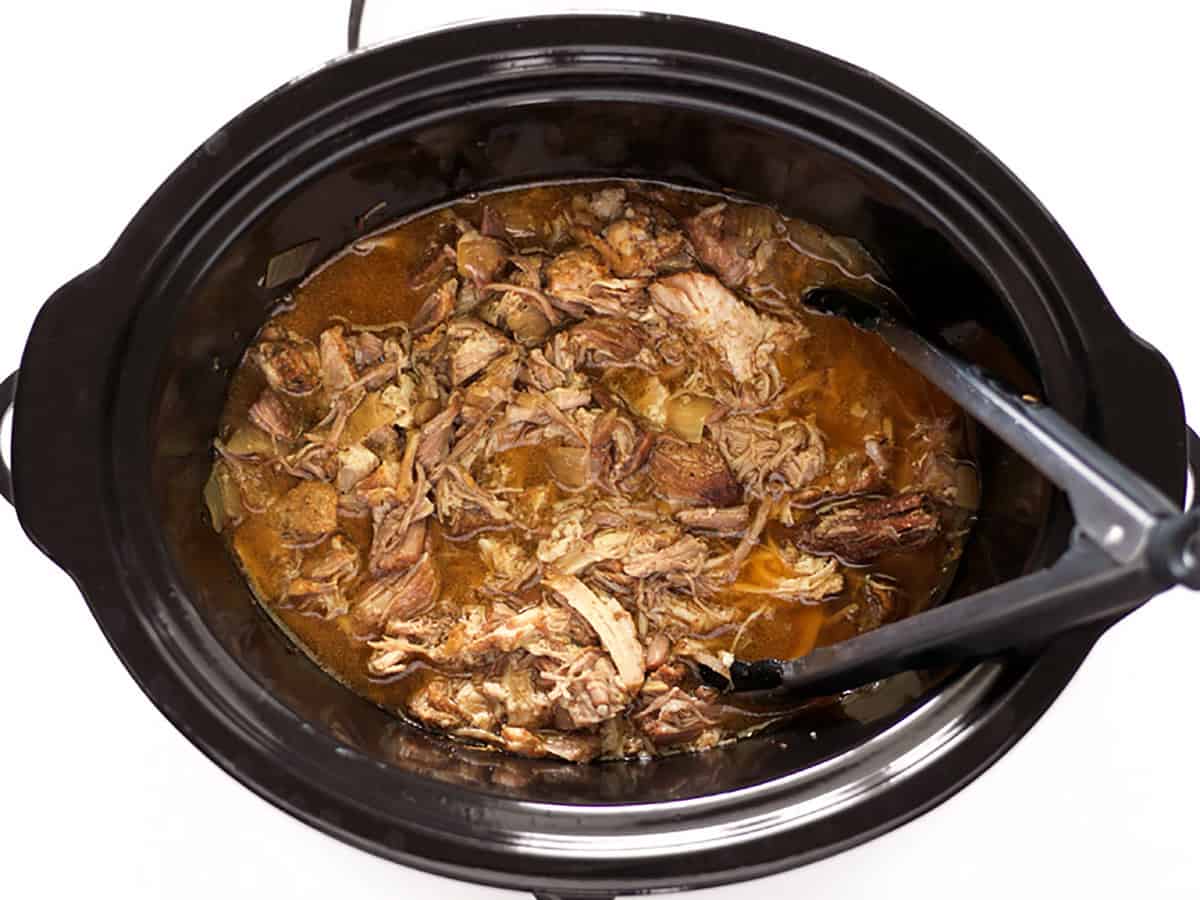 Shredded carnitas in the slow cooker with a pair of tongs.