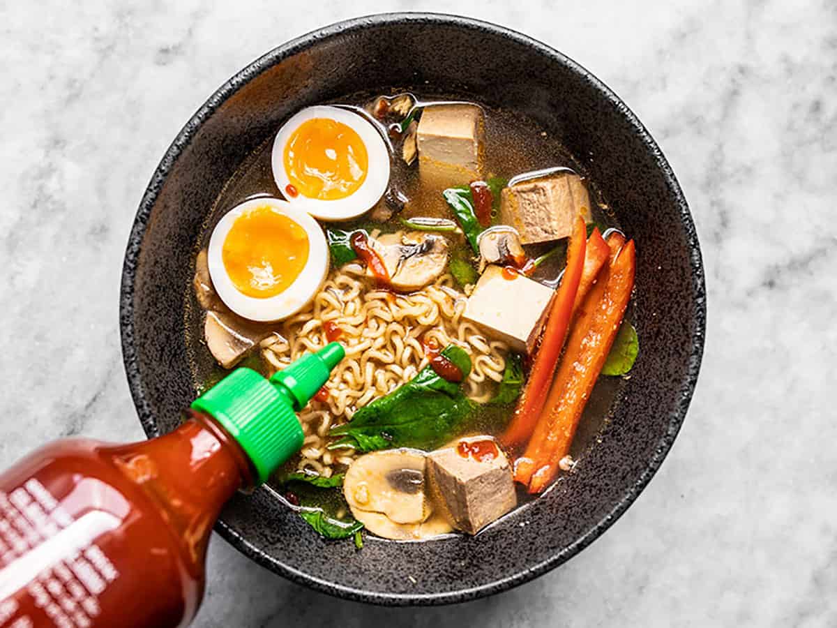 Sriracha being drizzled over a bowl of instant ramen with other ingredients added. 