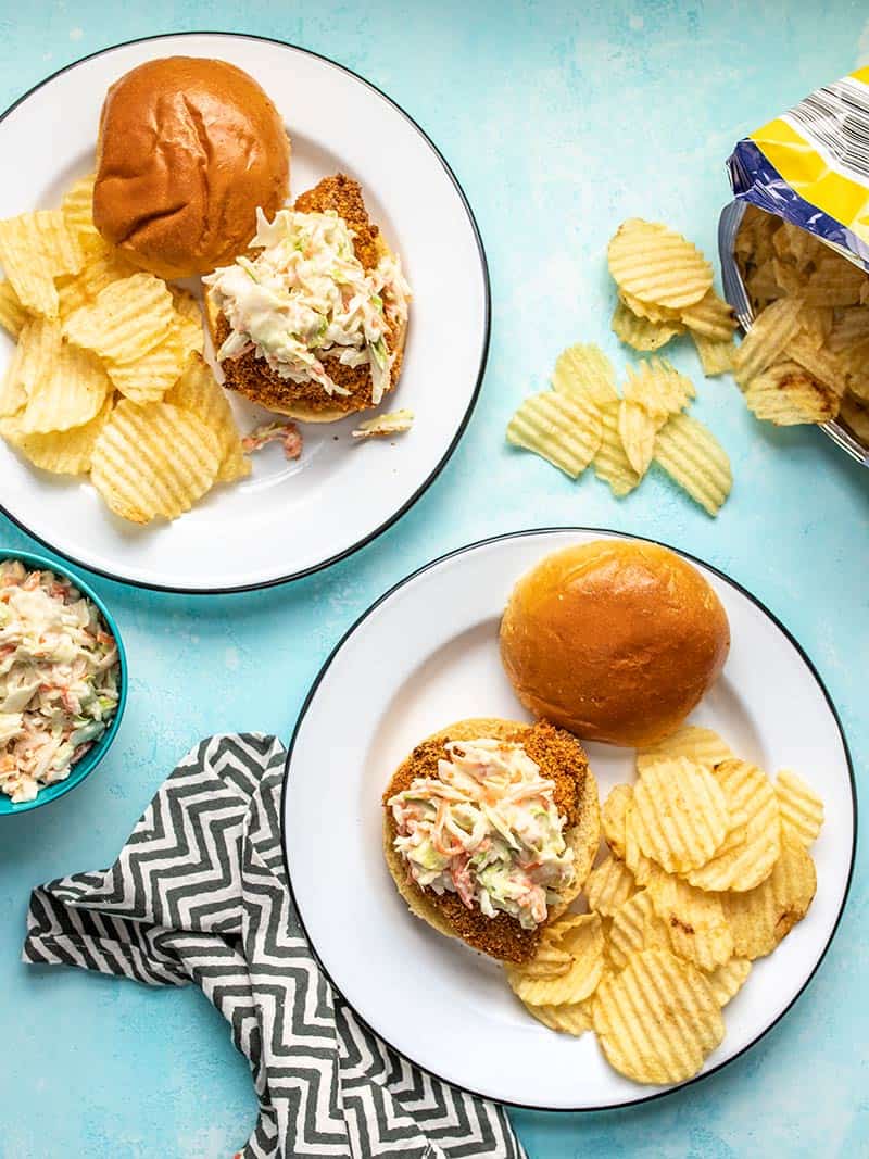 Overhead view of two plates with Baked Spicy Chicken Sandwiches with honey yogurt slaw and crinkle potato chips on a blue background. 
