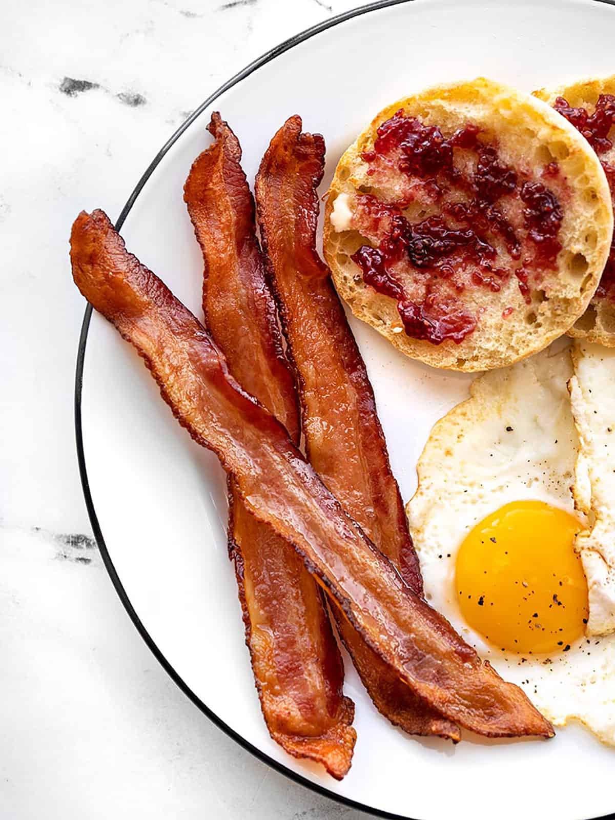 Close up of crispy oven baked bacon on a plate with a fried egg and English muffin.