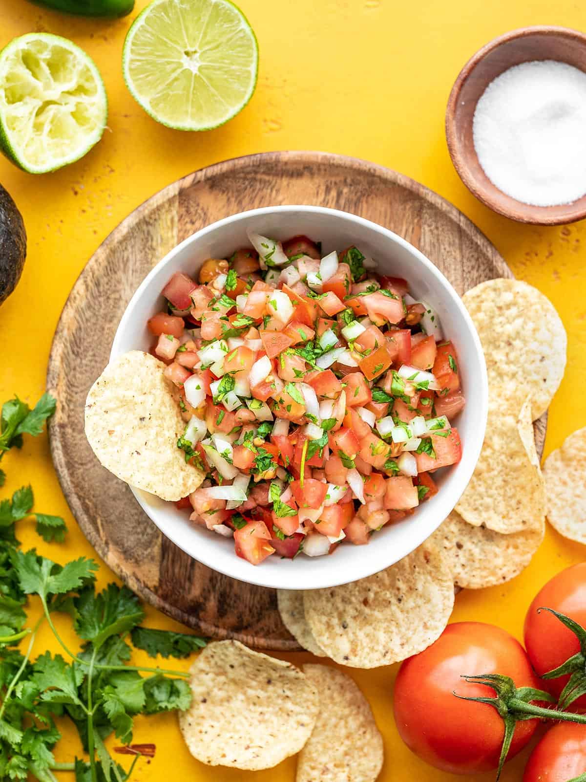 Overhead view of a bowl of pico de gallow with tortilla chips, salt, lime, and cilantro around it.