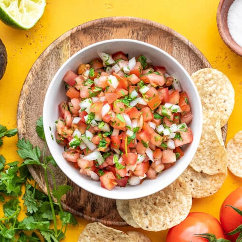 Overhead view of a bowl full of pico de gallo against a yellow background with ingredients and chips all around.