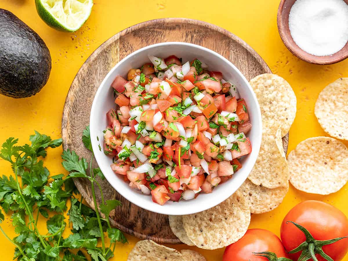 Overhead view of a bowl full of pico de gallo against a yellow background with ingredients and chips all around.