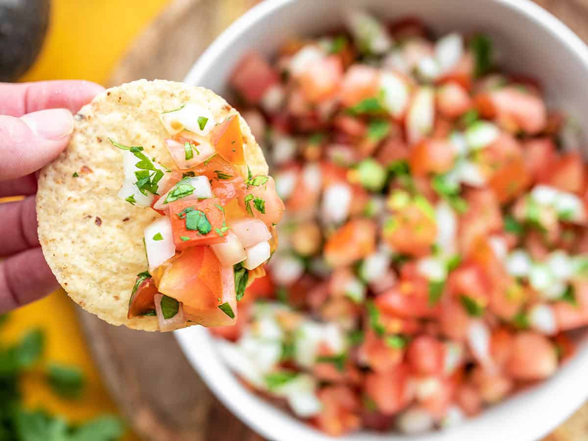 Close up of a tortilla chip with pico de gallo on it, the bowl of pico behind it.