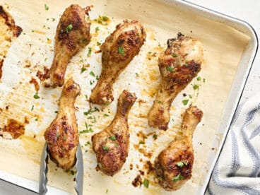 Overhead view of baked chicken drumsticks on a baking sheet, with one being removed with tongs.