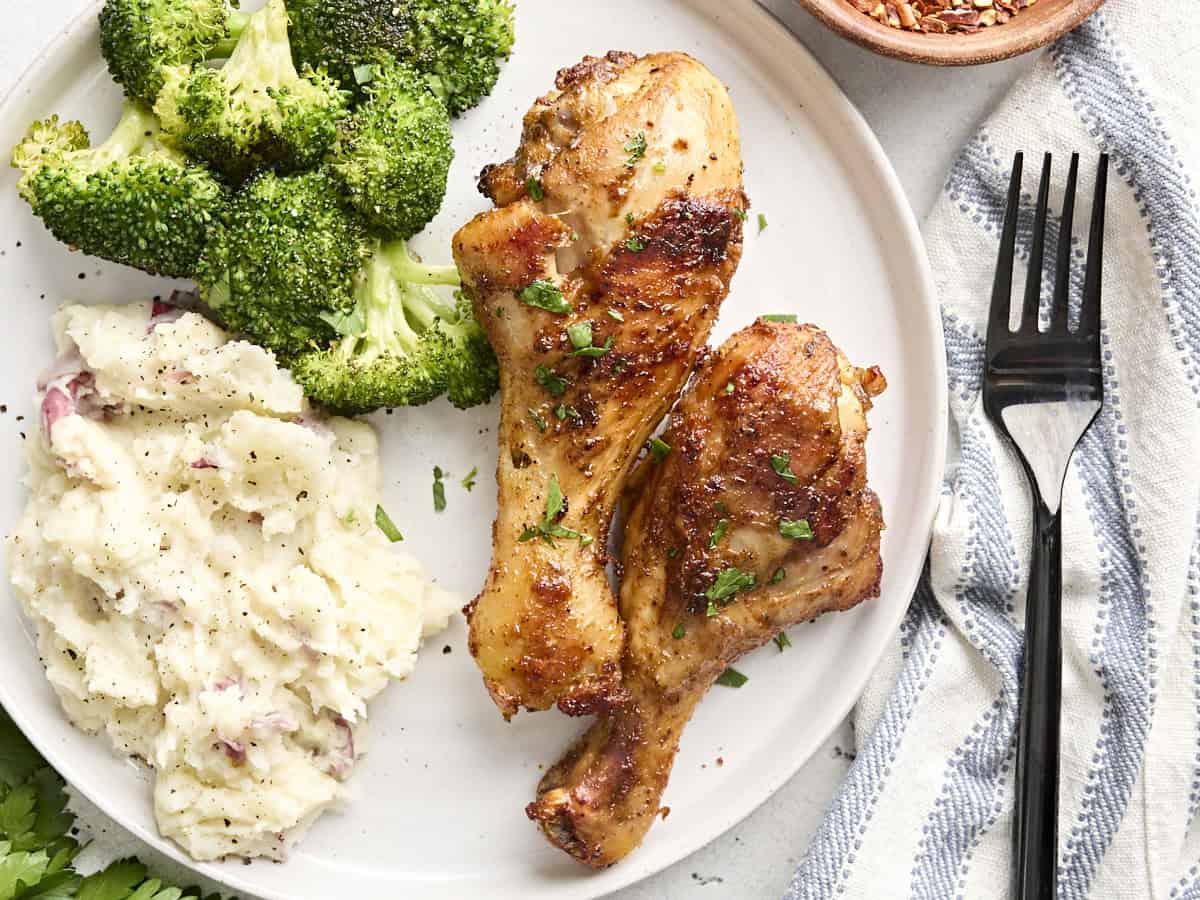 Overhead close up view of two baked chicken drumsticks on a plate with broccoli and mashed potatoes.