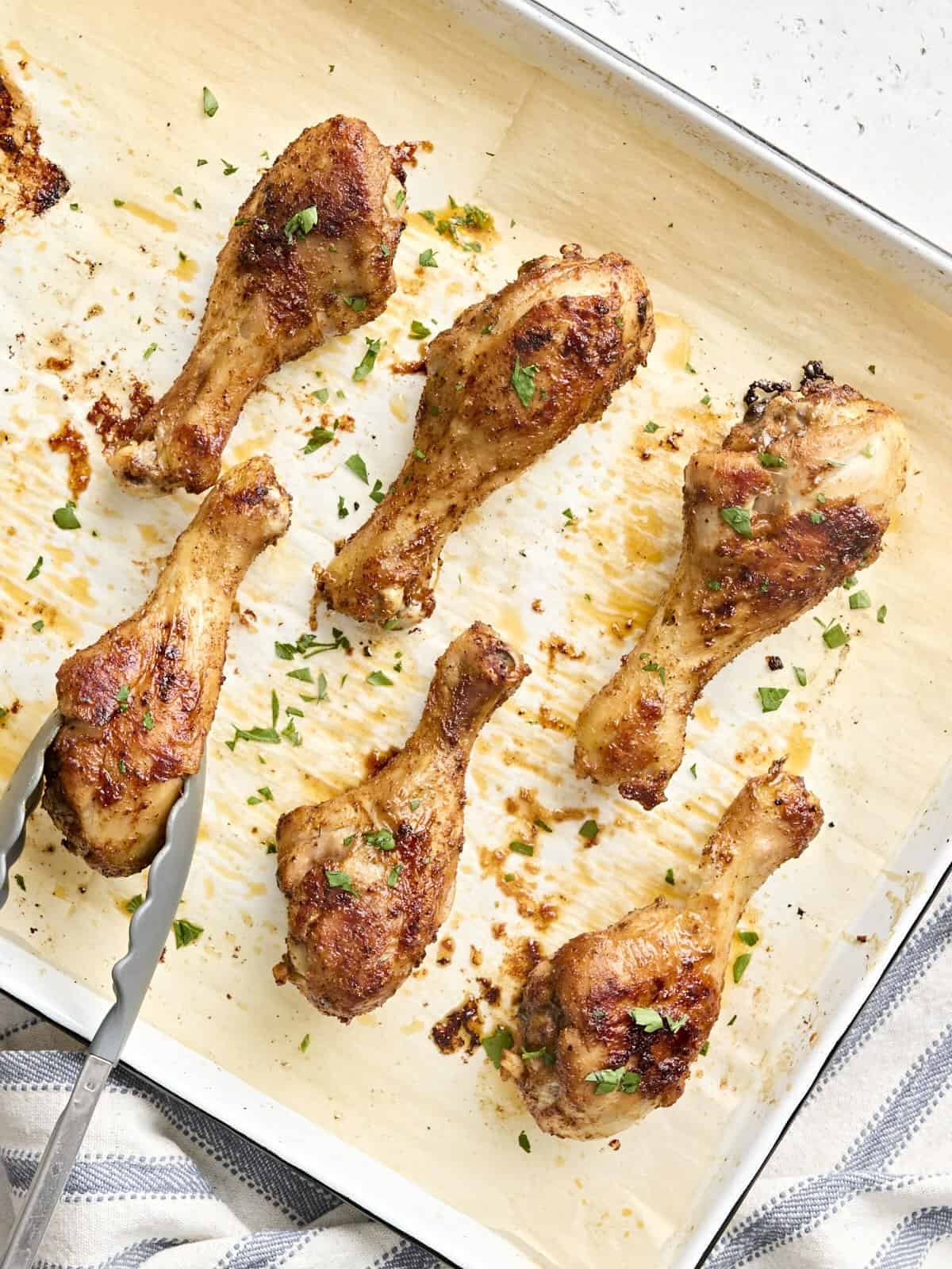 Overhead view of baked chicken drumsticks on a baking sheet, with one being removed with tongs.