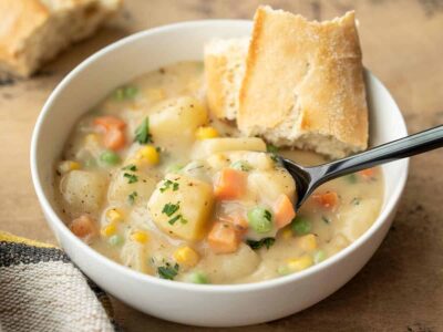 Side view of a bowl of vegetables with gravy with a piece of bread and a spoon.