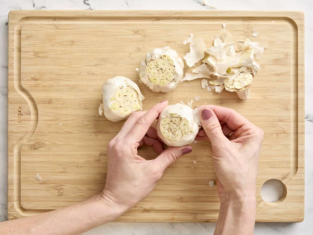 Hands peeling some of the skin off a head of garlic.