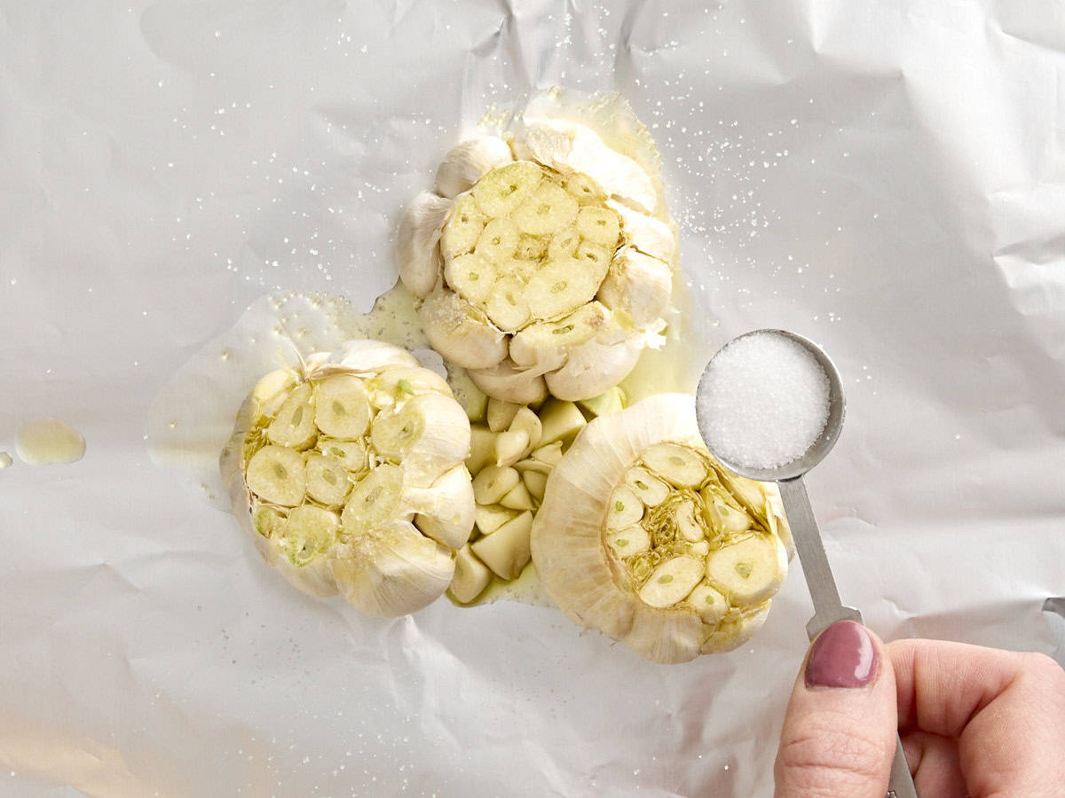 Salt being sprinkled over three heads of garlic on a piece of foil.