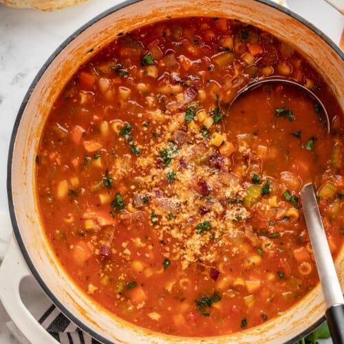 Dutch oven filled with finished Pasta e Fagioli soup topped with chopped parsley and parmesan cheese, with a ladle tucked into the right side of the soup.