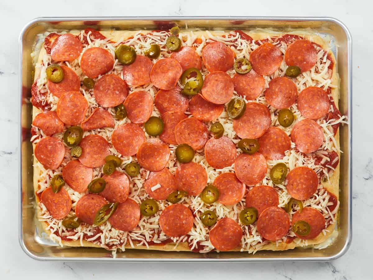 A par-baked pizza crust on a rimmed baking tray that's topped with pizza sauce, shredded mozzarella cheese, parmesan cheese, pepperonis and pickled jalapeño slices that needs to be baked again.