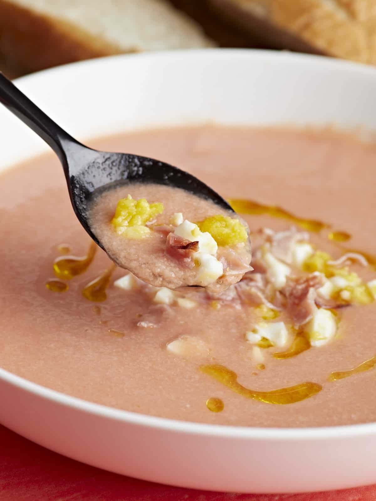 Side shot of a white bowl of Salmorejo garnished with a drizzle of olive oil, chopped hard-boiled eggs, and crispy ham crumbles with a black spoon coming out of it.