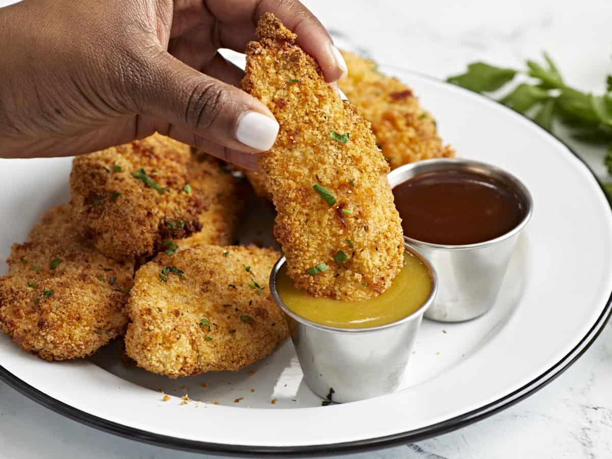 Side view of air fryer chicken tenders on a serving plate with one being dipped in honey mustard sauce.