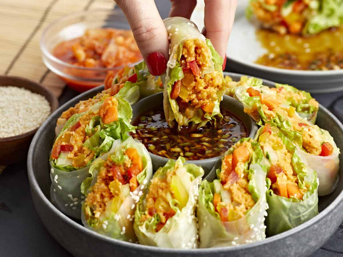 dipping a kimchi spring roll into a vat of sauce in the center of a platter.