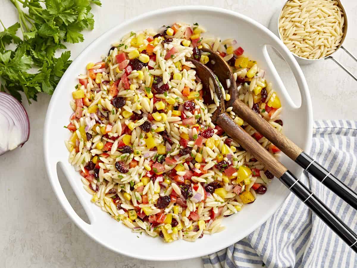 Overhead view of orzo pasta salad in a large white serving bowl with wooden serving spoons on the side.