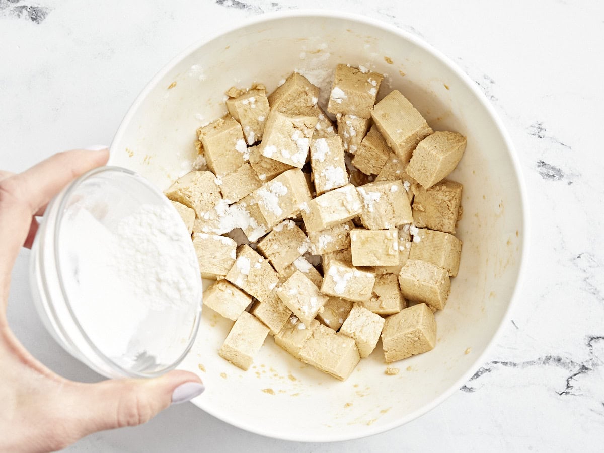 pouring cornstarch over diced tofu in a white bowl.
