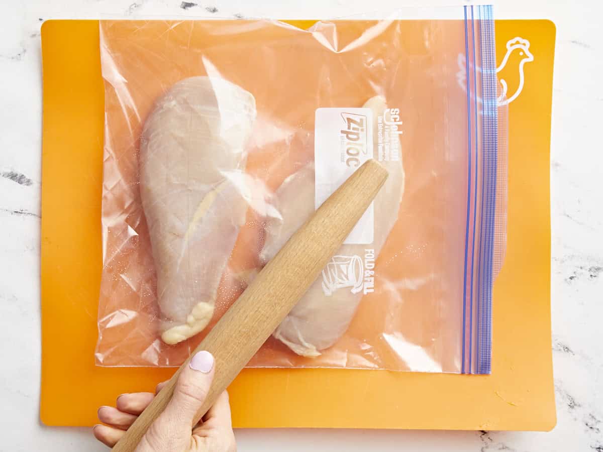 pounding chicken breasts in a ziplock bag with a rolling pin on an orange cutting board.