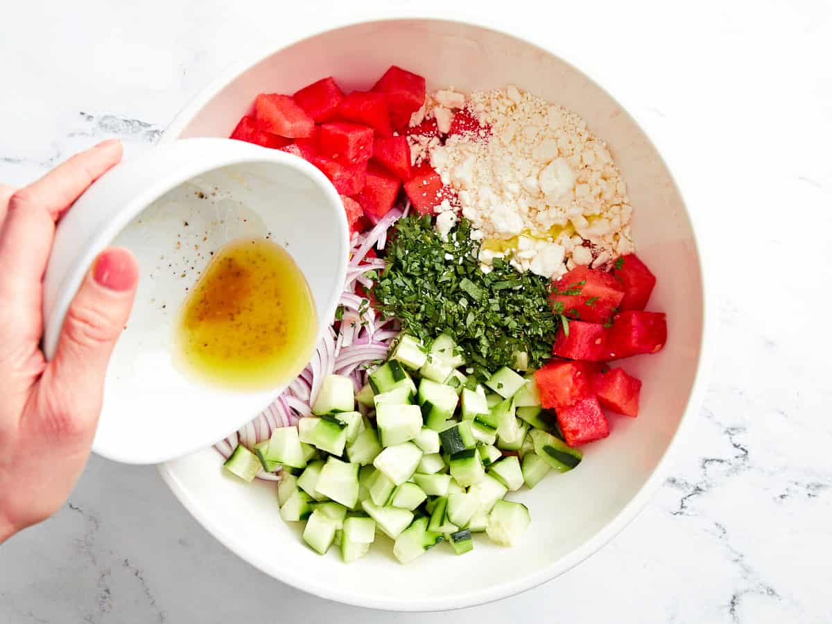 pouring dressing over watermelon and feta salad in a white bowl.