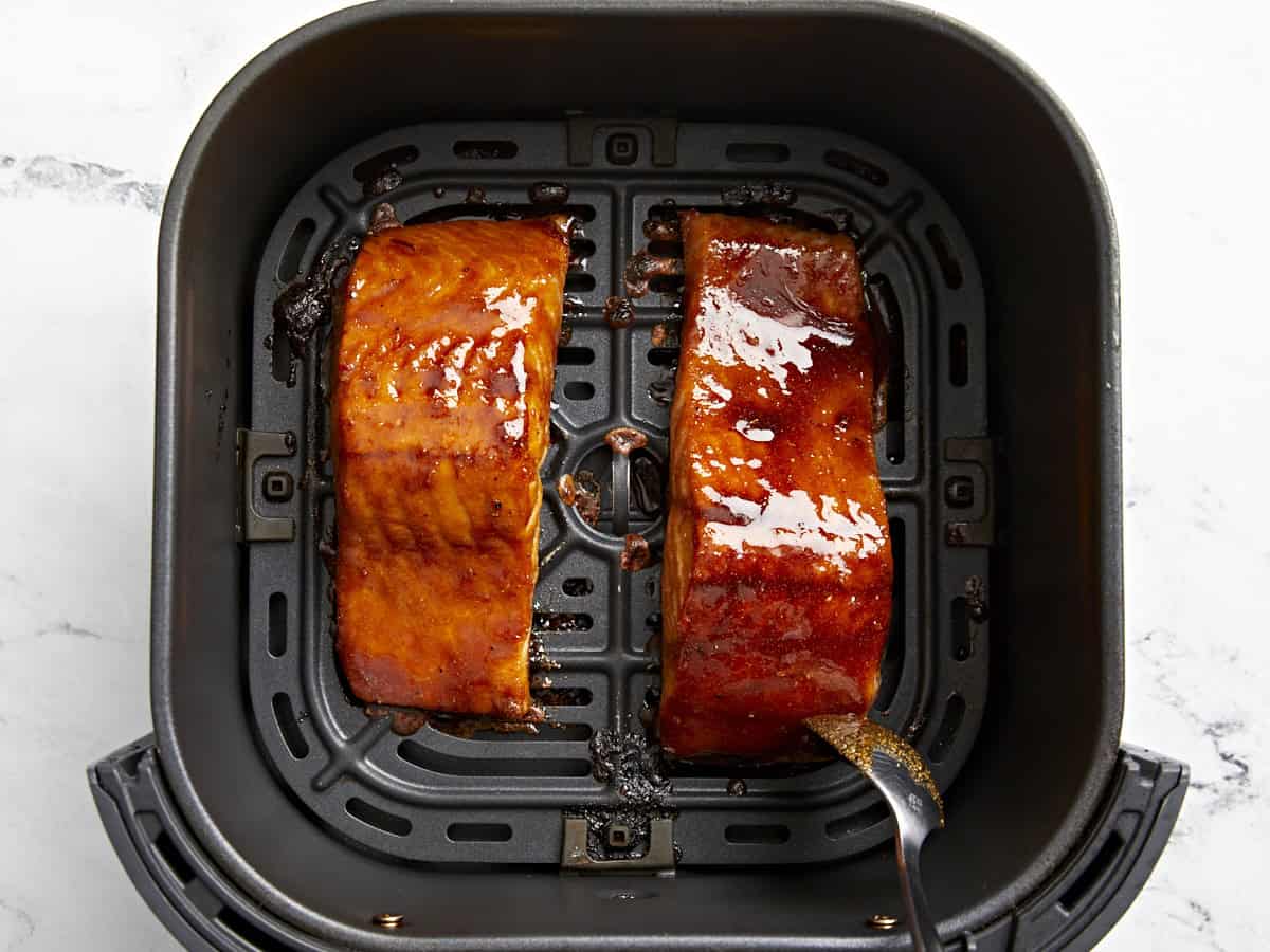 spooning marinade over partially cooked salmon filets in an air fryer basket.