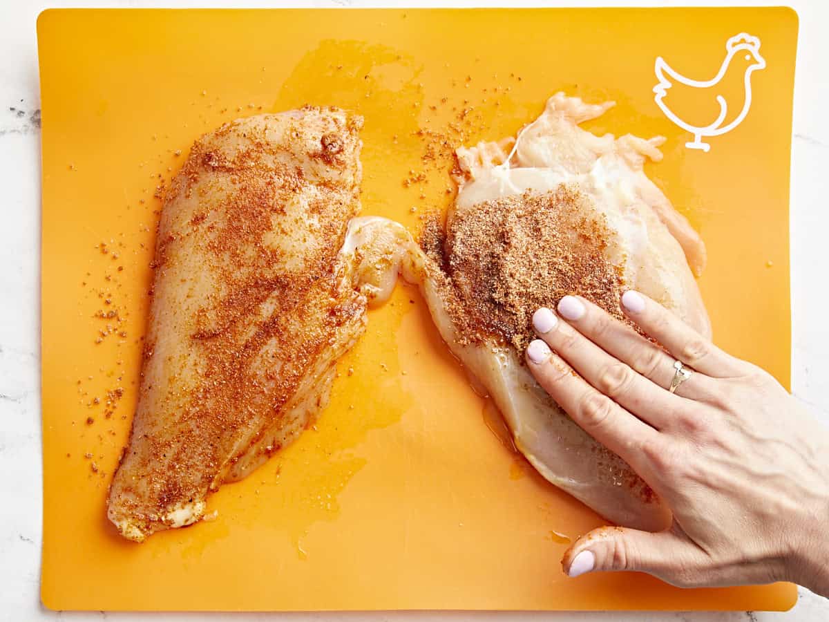 pressing seasoning onto chicken breasts on an orange cutting board.