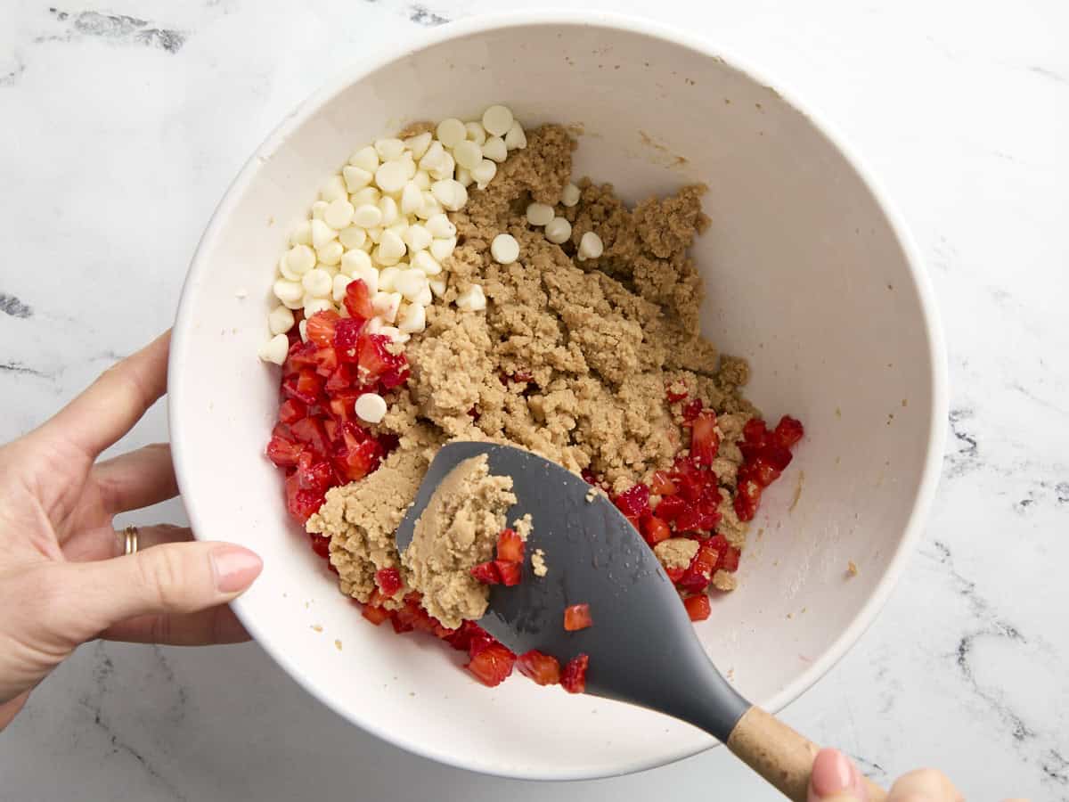 folding strawberry pieces and white chocolate chips into cookie dough in a white bowl with a rubber spatula.