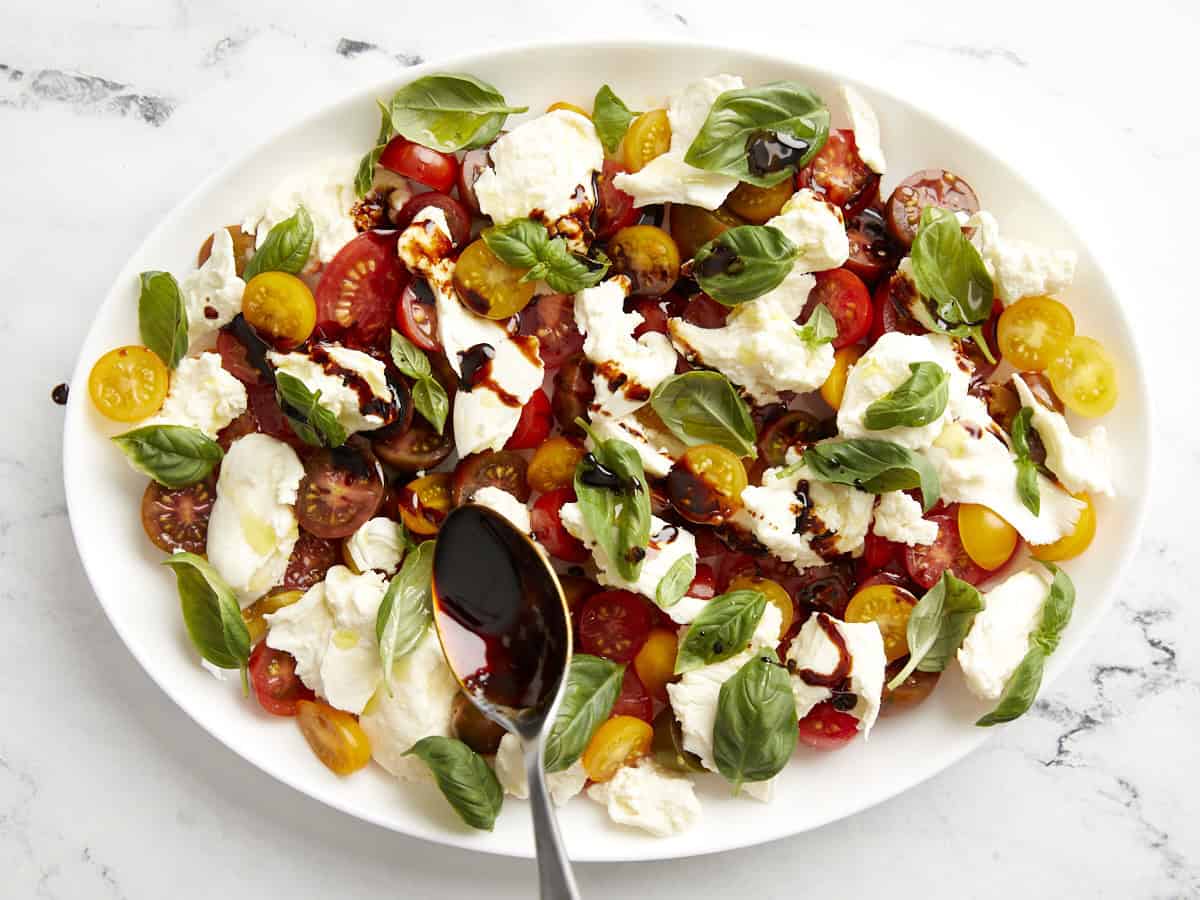 drizzling balsamic glaze over caprese salad on a white serving plate with a spoon.