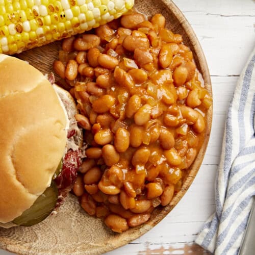 overhead view of a serving of baked beans on a plate with a sandwich and corn on the cob.