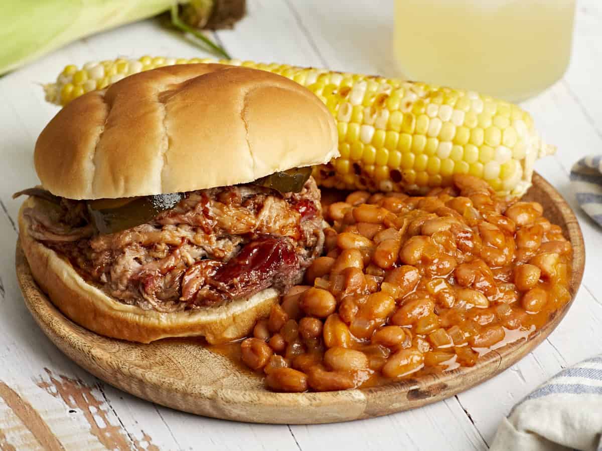a serving of baked beans on a plate with a pulled pork sandwich and corn on the cob.