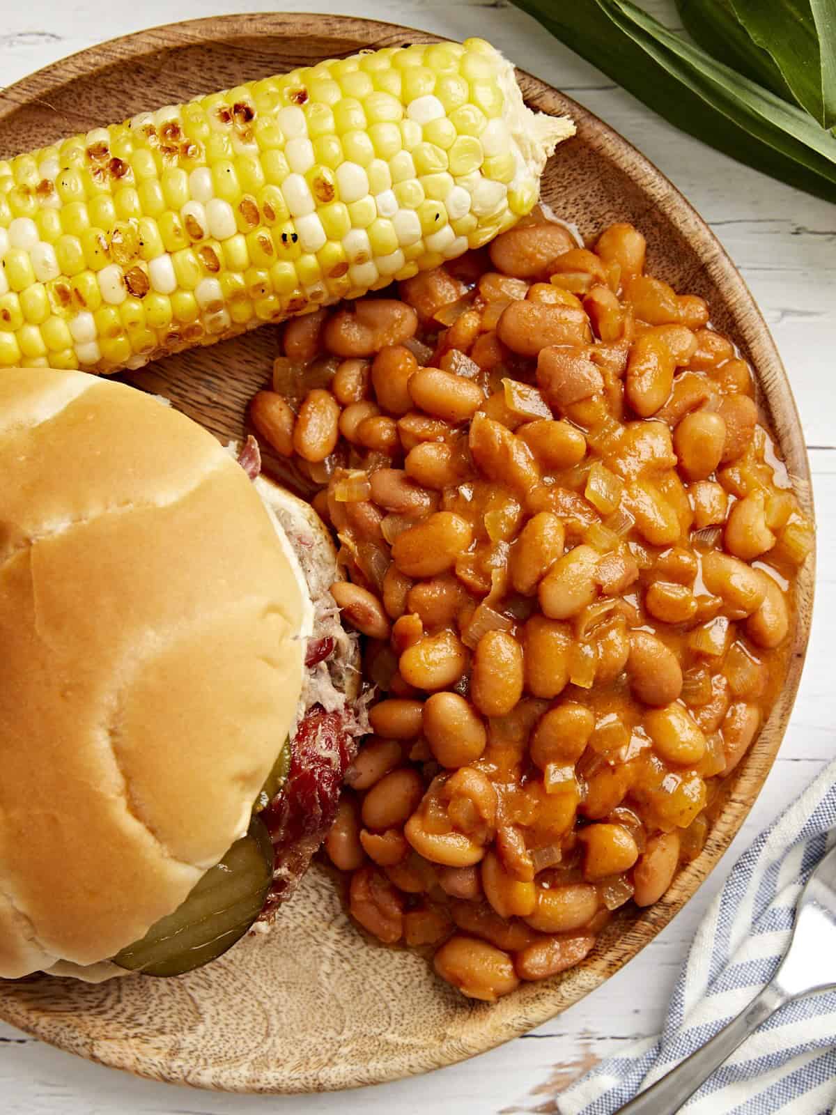 overhead view of a serving of baked beans on a plate with a sandwich and corn on the cob.