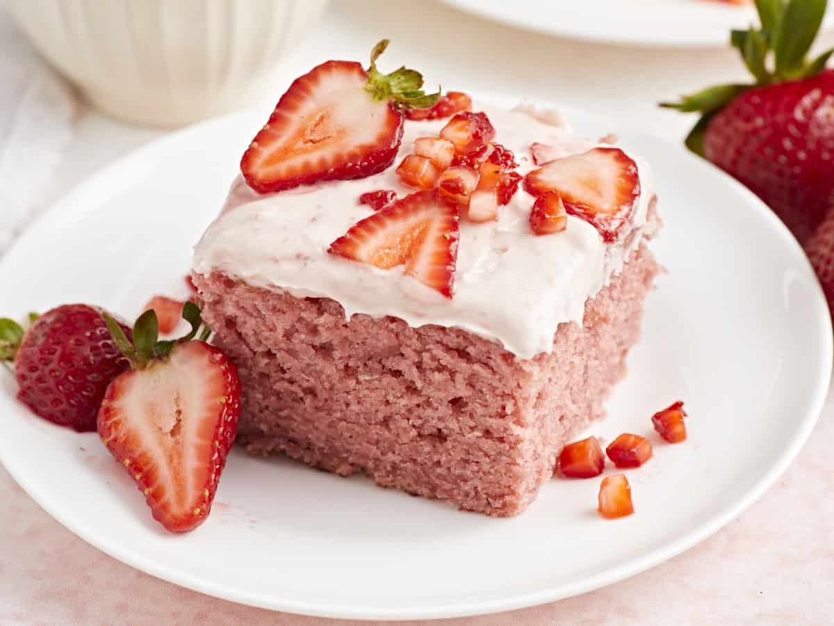 Close up view of strawberry sheet cake on a white plate with fresh strawberries on top.