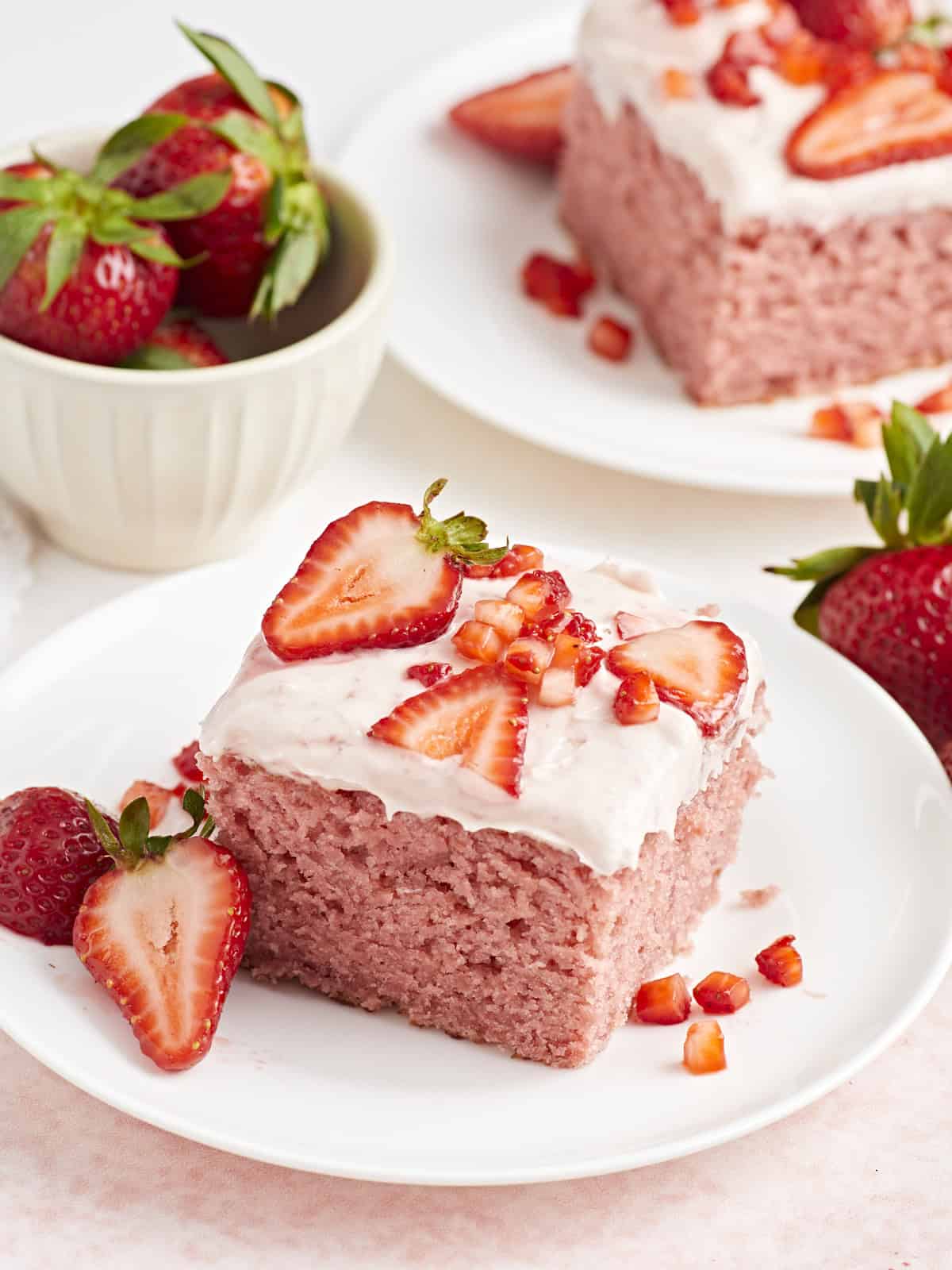 Side view of a slice of strawberry sheet cake on a small white plate with fresh strawberries on the side.