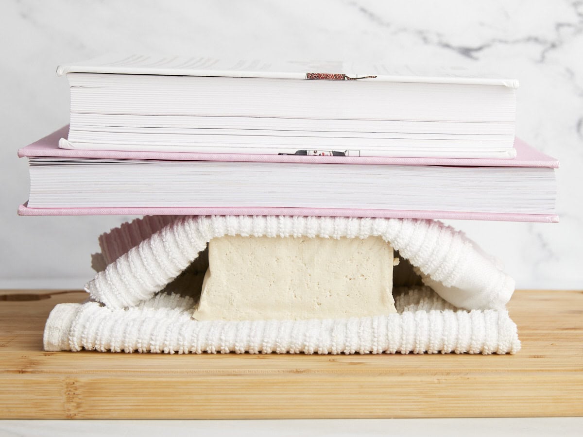 A block of tofu being pressed between two towels and cook books.