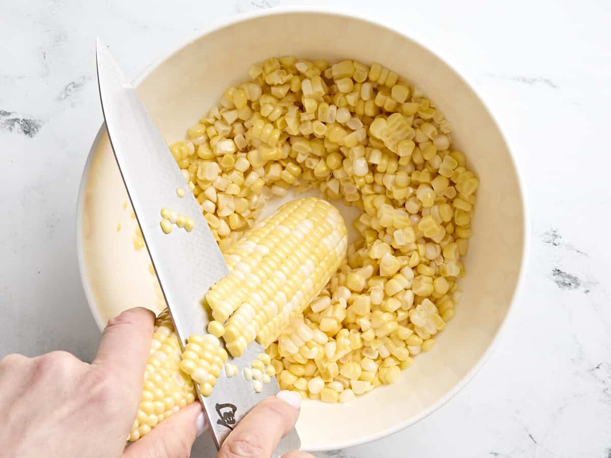 A knife slicing kernels off a corn cob into a bowl.