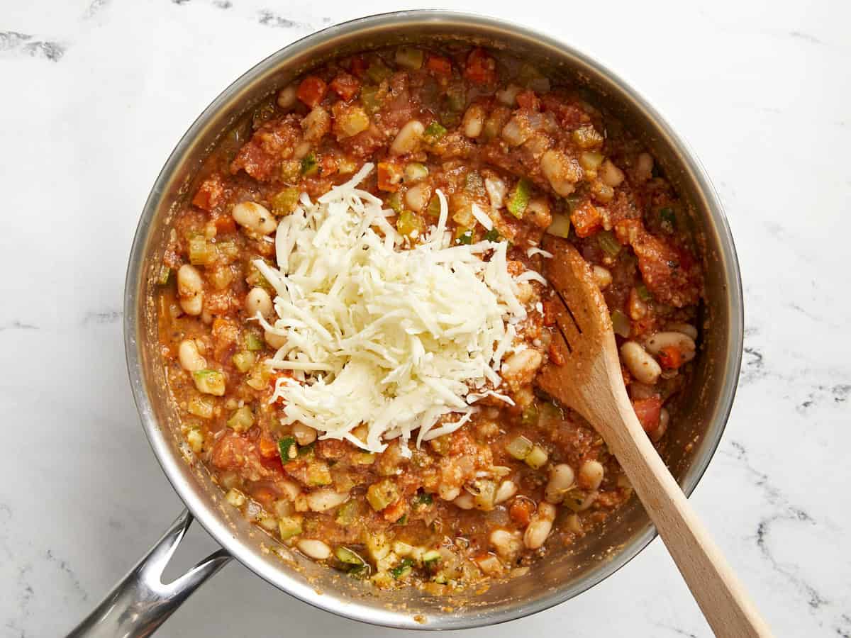 Filling for tuscan white bean stuffed tomatoes in a saucepan topped with shredded cheese.