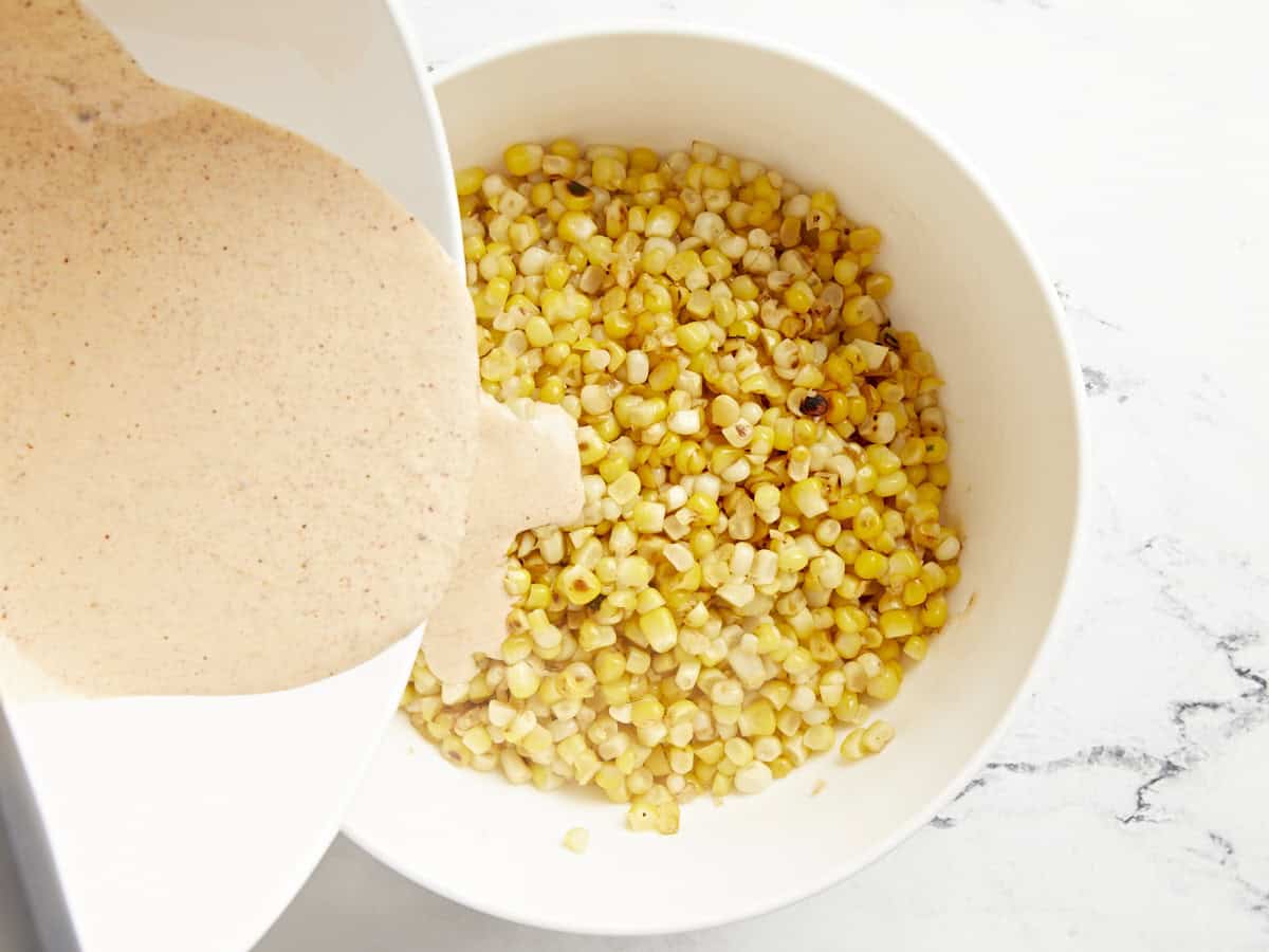 Mexican street corn salad dressing being poured into a bowl of charred corn kernels.