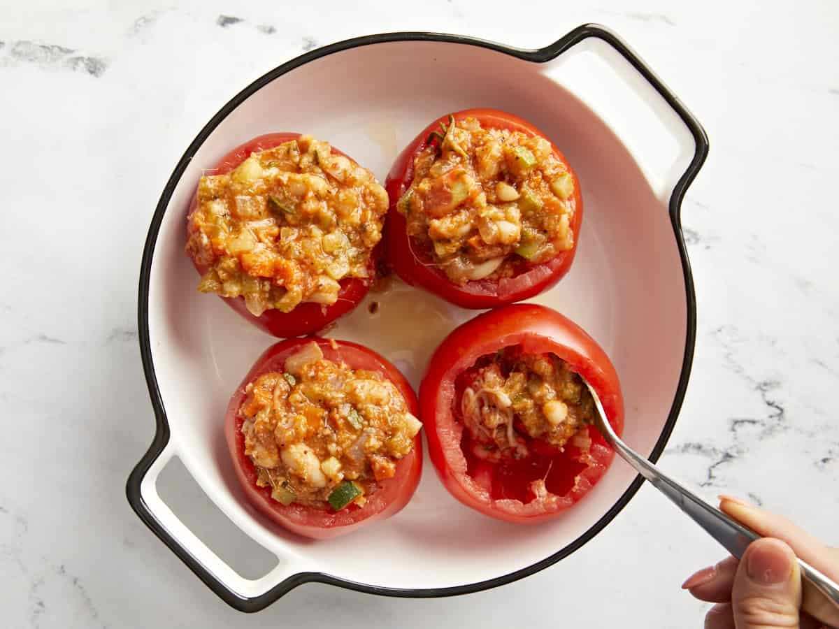 A spoon adding the tuscan white bean filling to hollowed out tomatoes in a baking dish.